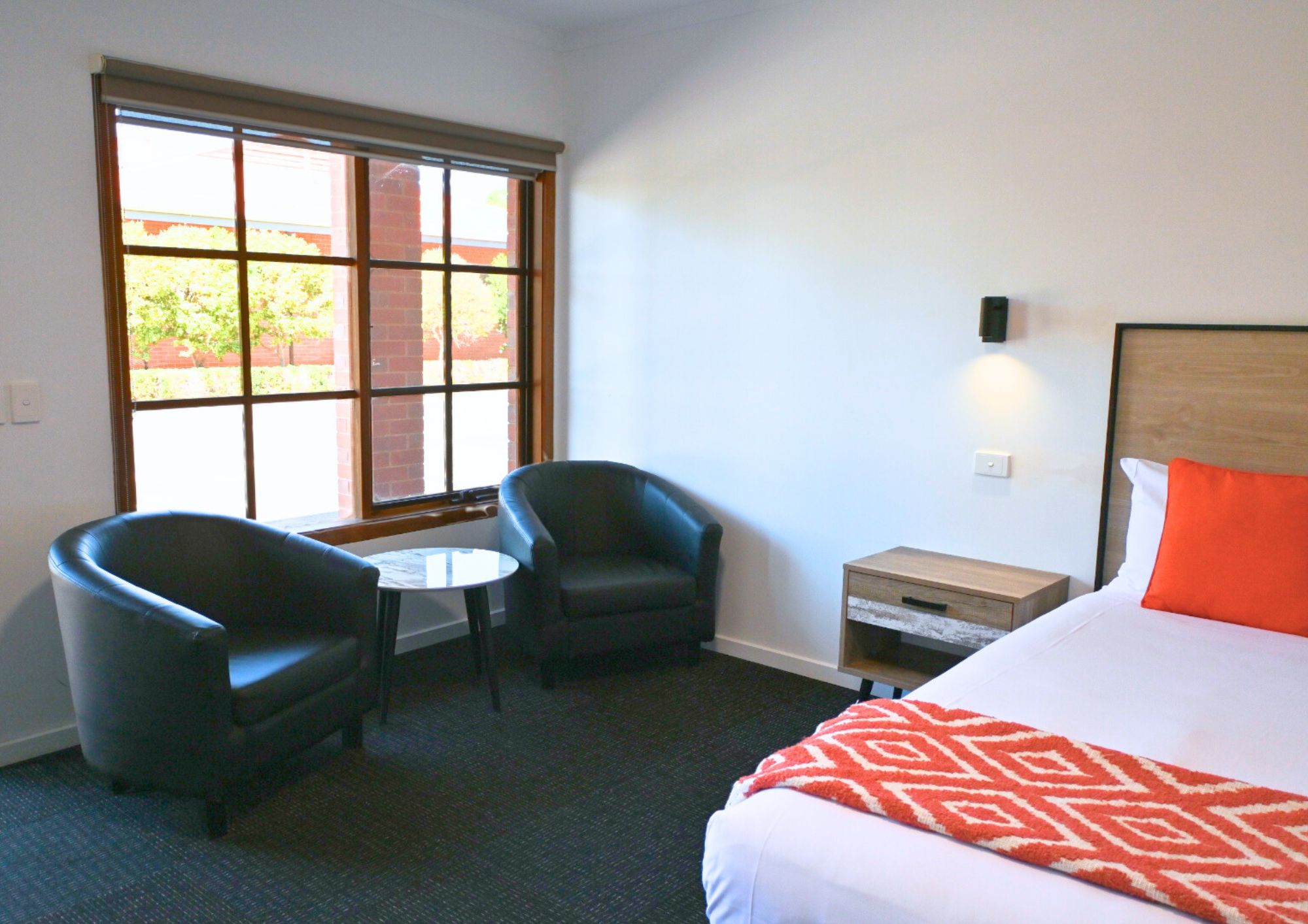 A hotel room with a bed, two armchairs, a small table, a nightstand, and a window with a view of greenery.