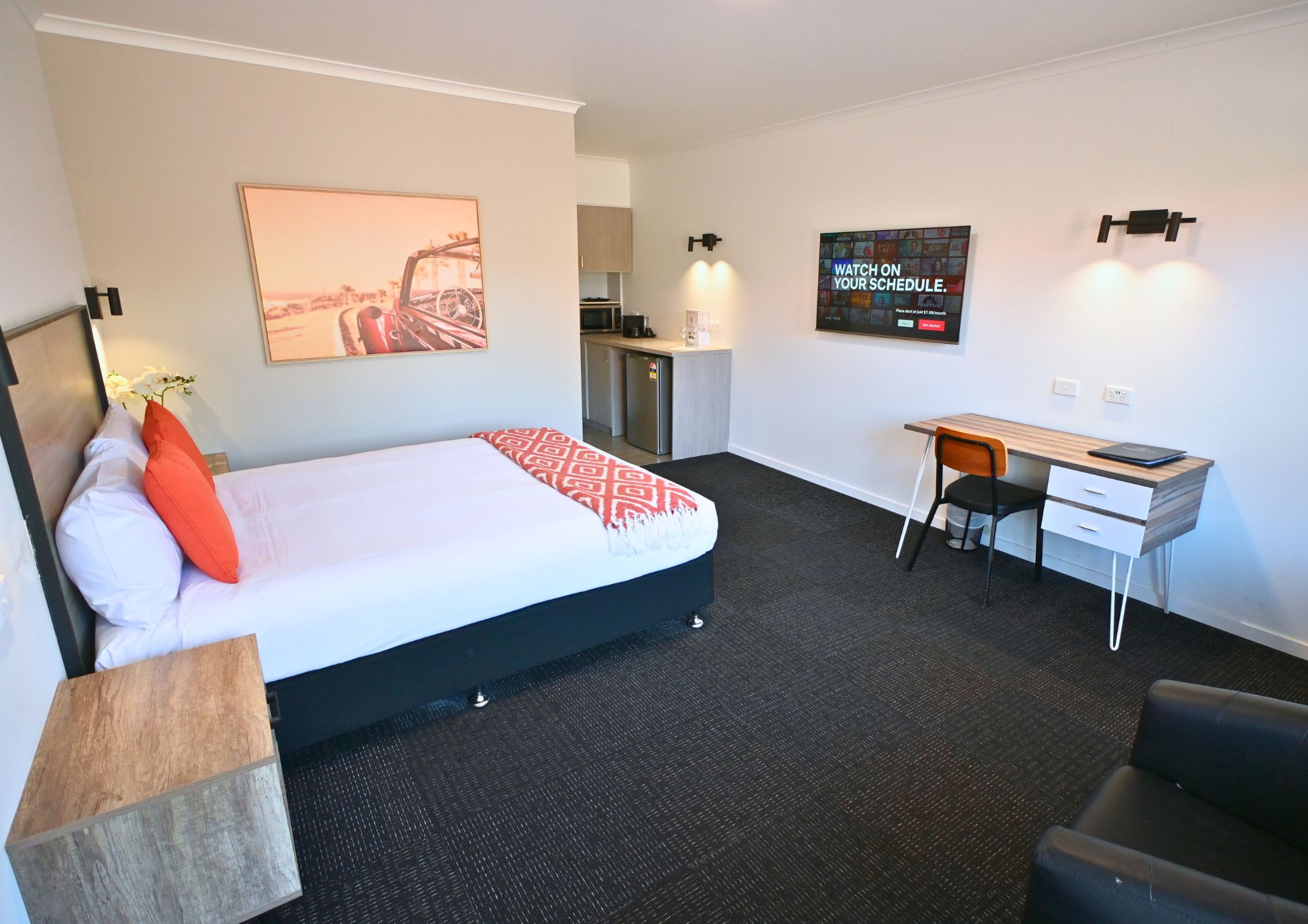 A hotel room with a bed, a desk, a chair, a TV, and a kitchen area.