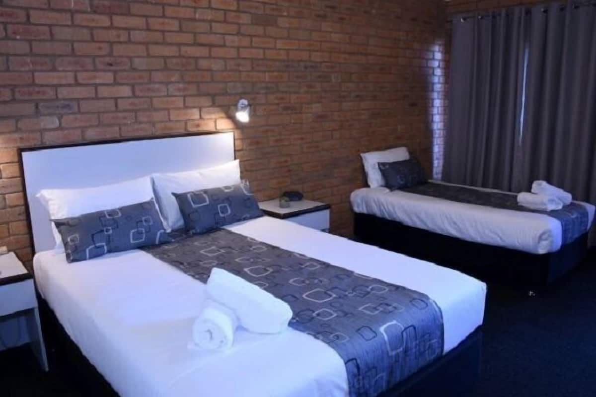 A hotel room with a brick wall, a large bed with a headboard, a smaller bed, a nightstand with a lamp, and a window with curtains.