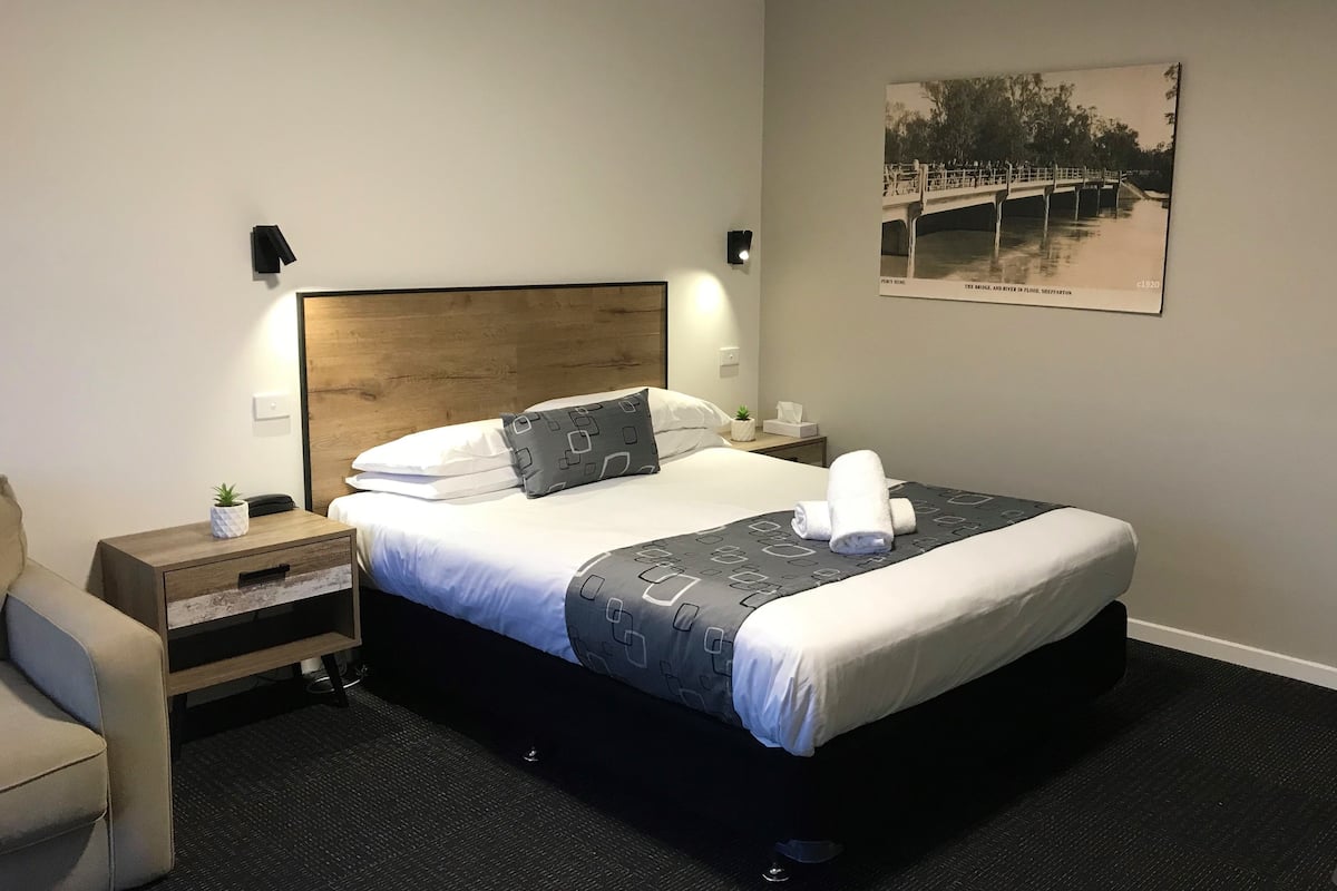 A neatly made bed with a wooden headboard, a bedside table with a plant, a wall-mounted lamp, and a framed picture on the wall.