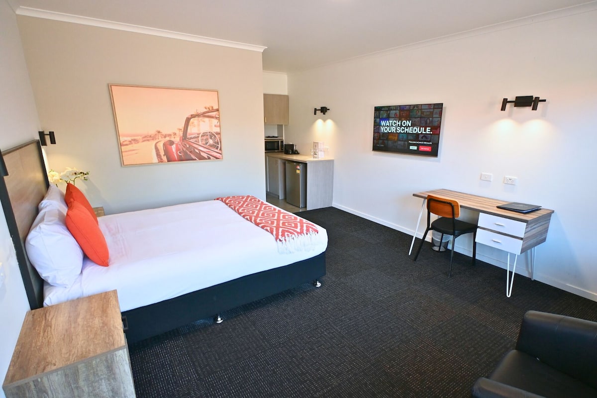 A hotel room with a bed, a desk, a chair, a TV, and a kitchen area.