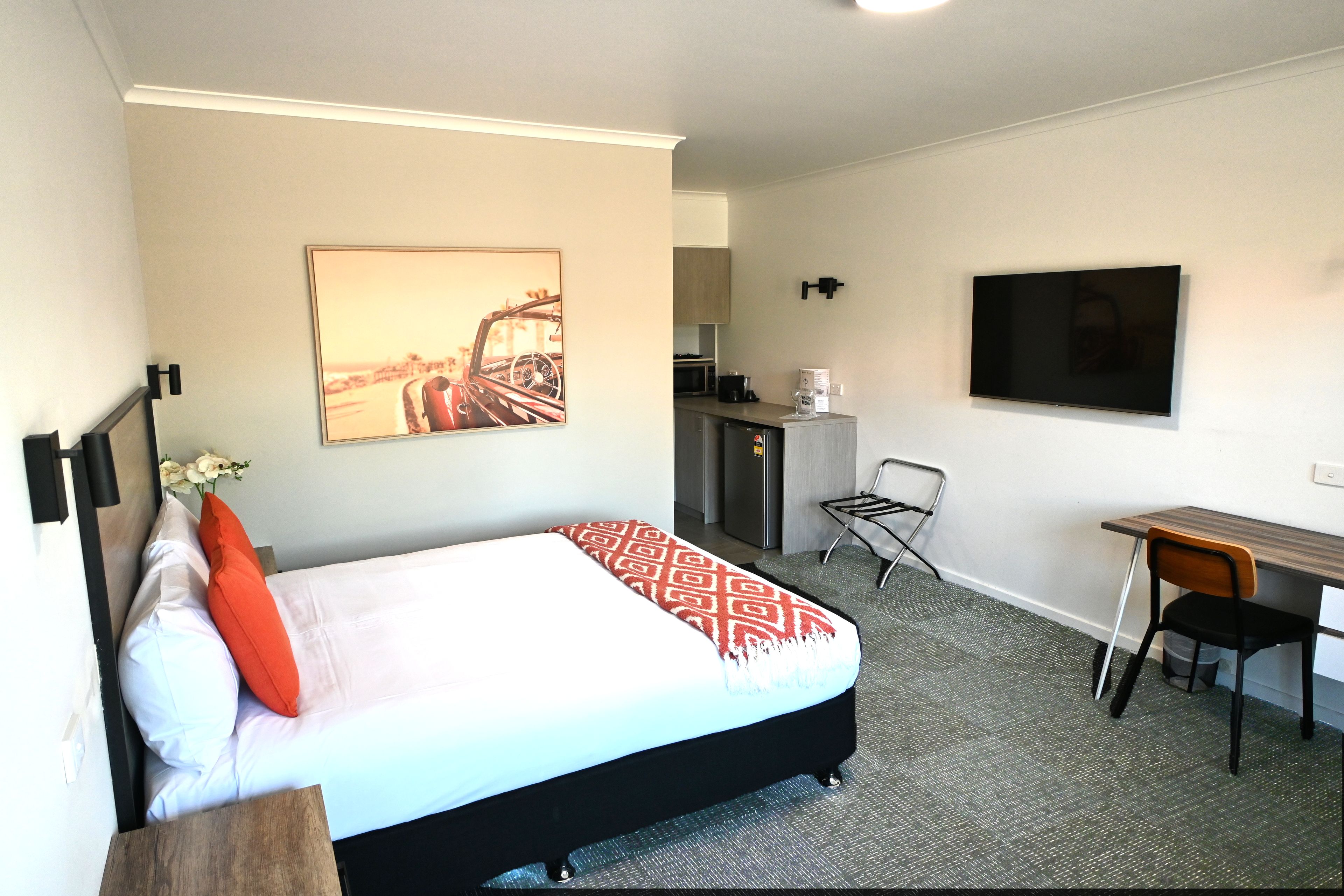 A hotel room with a bed, a desk, a chair, a television, a kitchen area, and a framed picture on the wall.