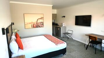 A hotel room with a bed, a desk, a chair, a television, a kitchen area, and a framed picture on the wall.
