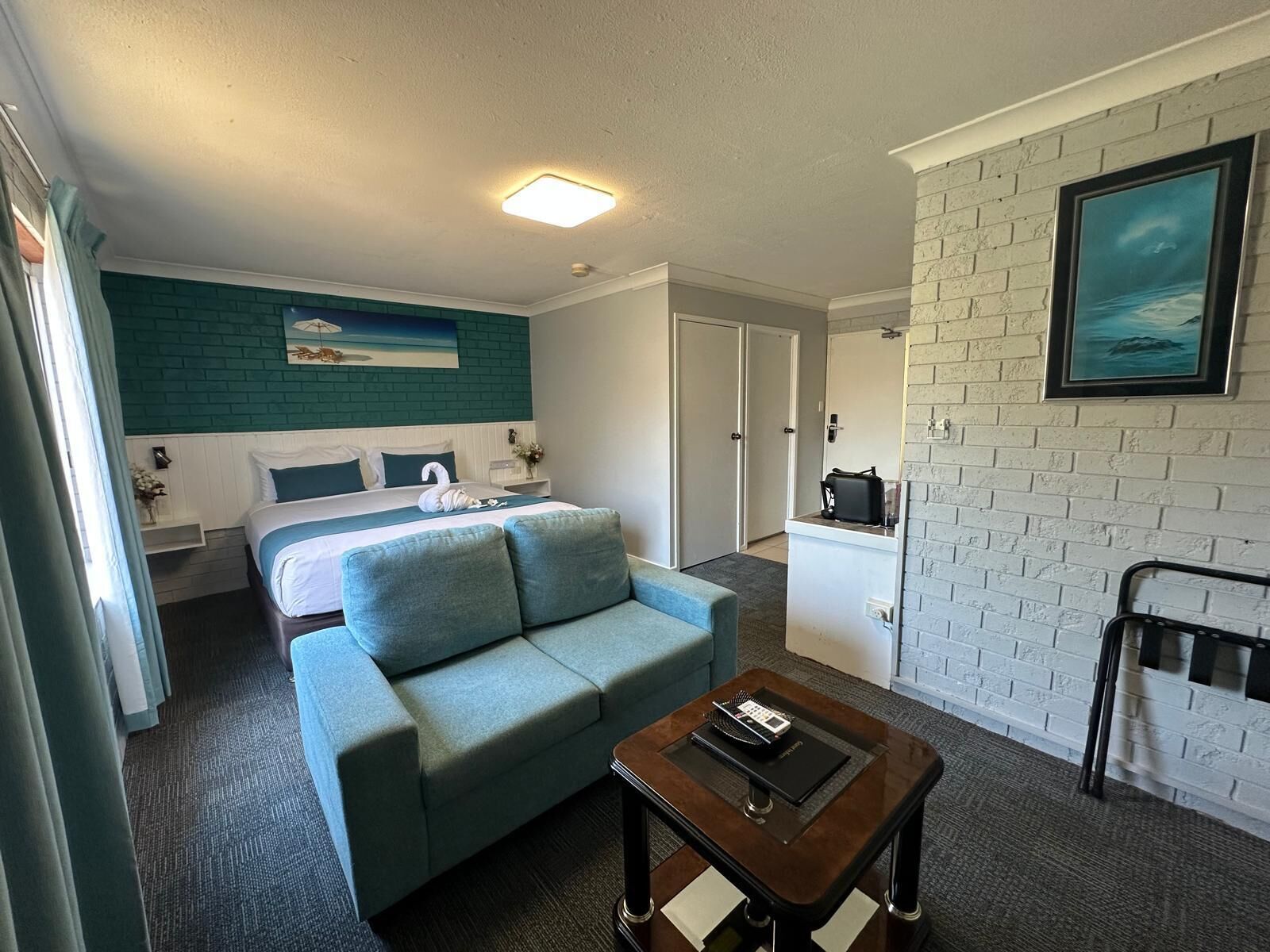 A hotel room with a bed, a blue sofa, a small table, a white brick wall, and a painting.