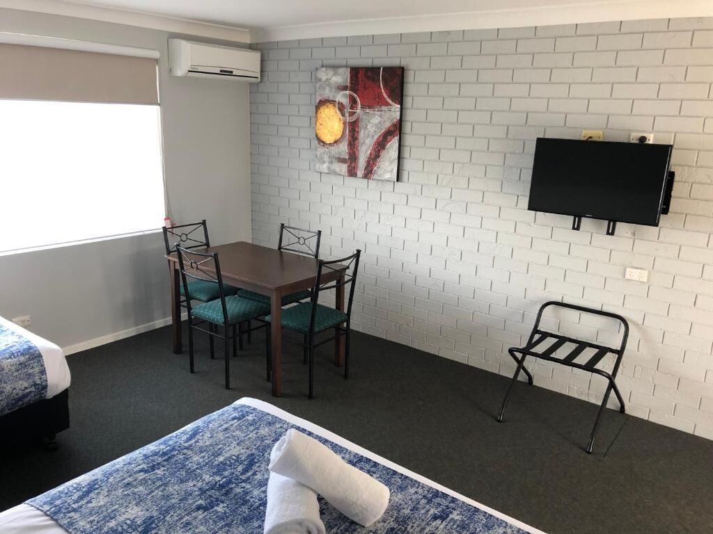 Quarto de hotel com parede de tijolos brancos, televisão de tela plana sobre uma mesa de madeira com quatro cadeiras, e duas camas com roupa de cama azul.