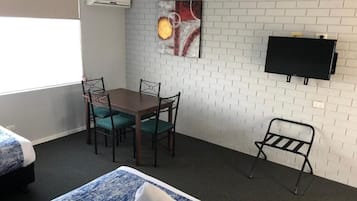 Habitación de hotel con pared de ladrillo blanco, un televisor de pantalla plana sobre una mesa de madera con cuatro sillas y dos camas con ropa de cama azul.