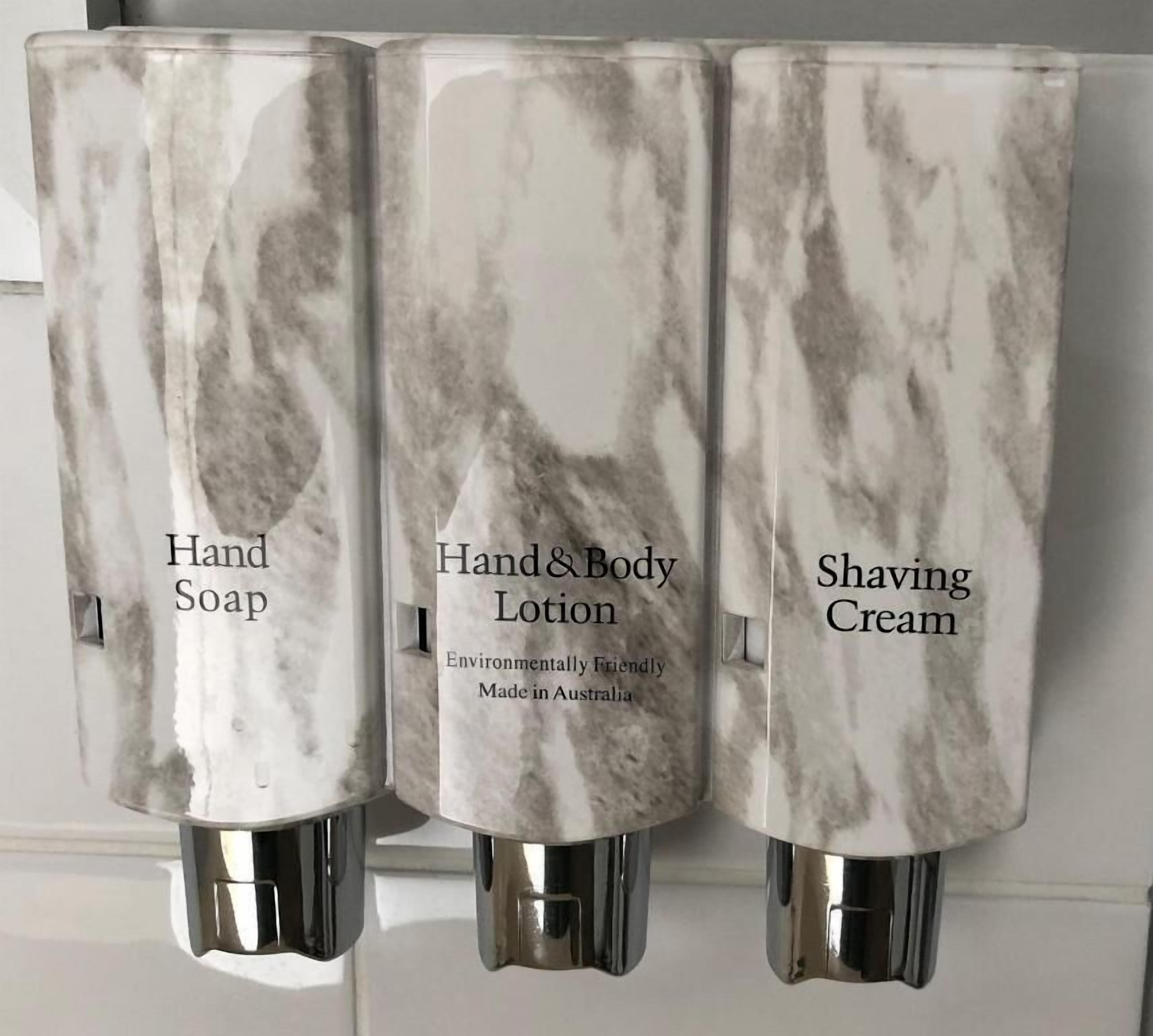 Tres dispensadores de baño etiquetados con diferentes productos: jabón de manos, loción corporal y crema de afeitar.