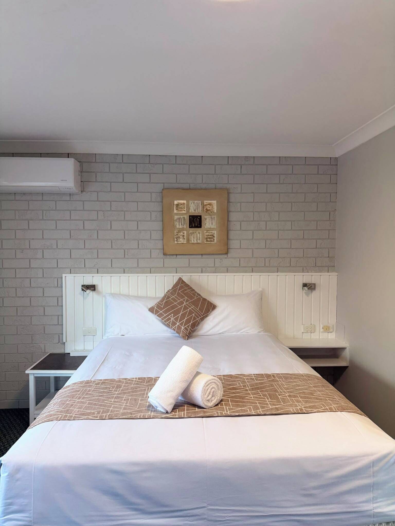 A hotel room with a bed, a white headboard, a brick-patterned wall, and a framed artwork above the bed.