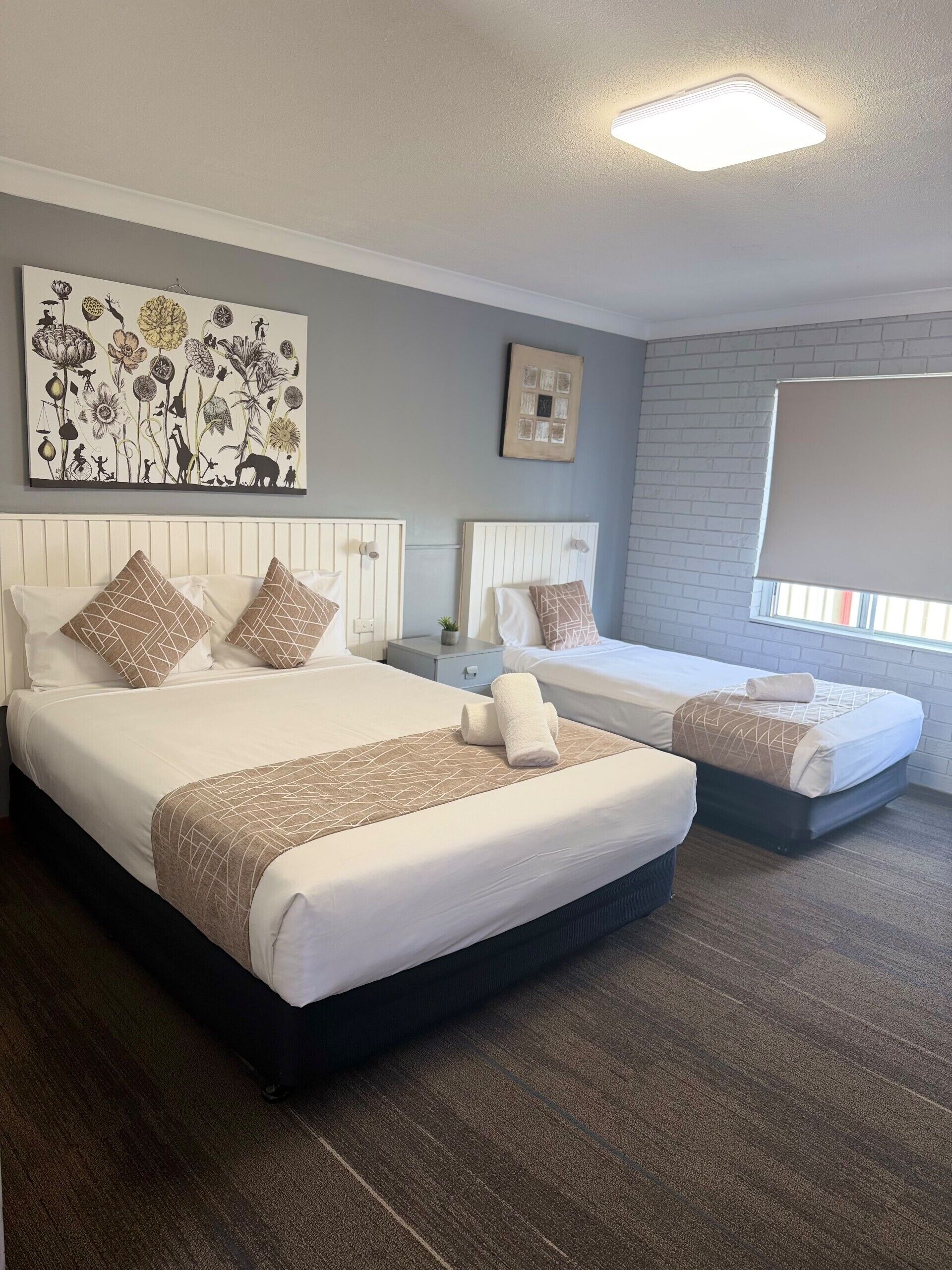 A hotel room with two beds, a large floral artwork, a small plant, and a window with a view.