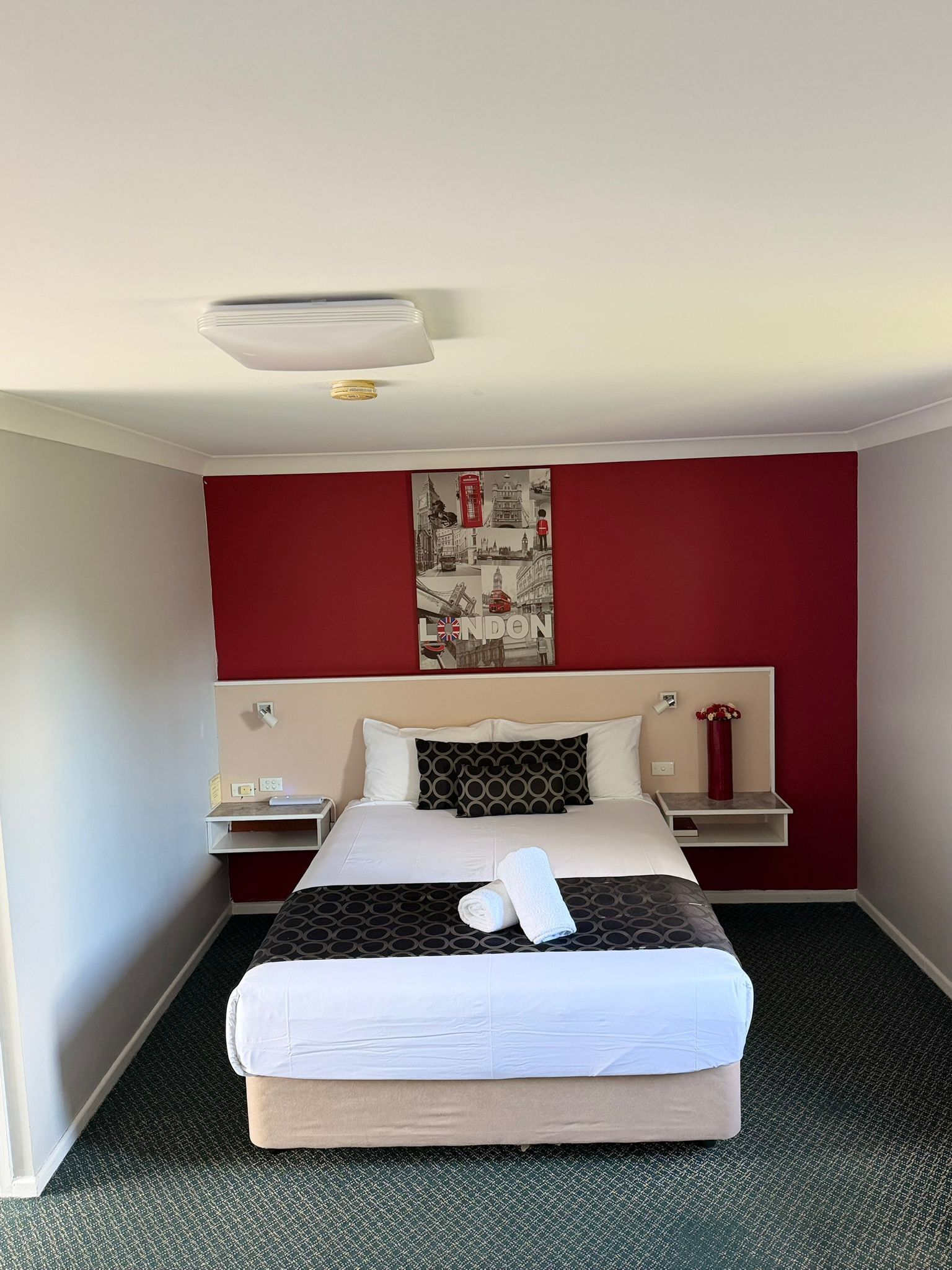 A hotel room with a bed, a nightstand, a red wall, and a London-themed artwork.