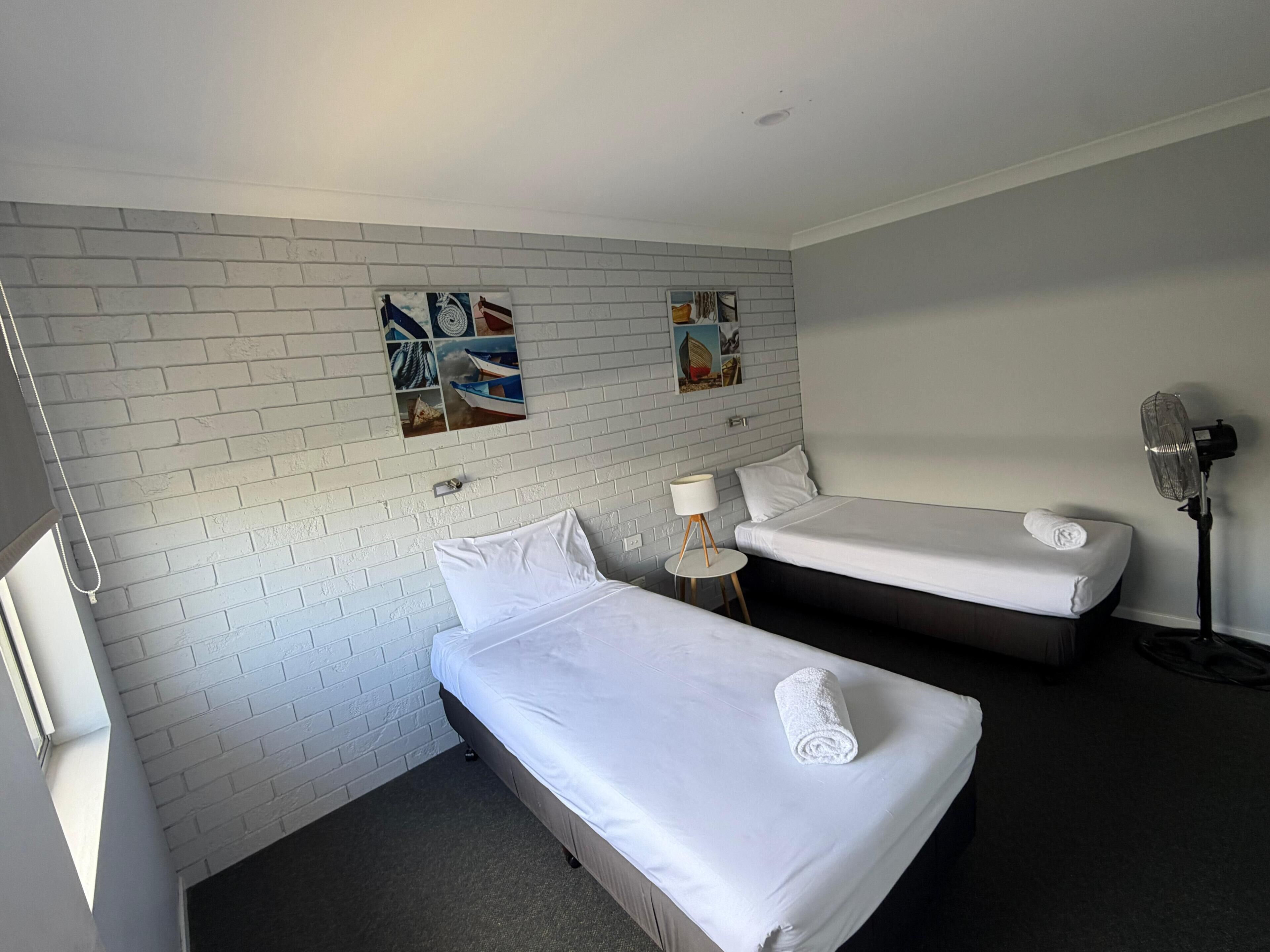 Une chambre d’hôtel avec deux lits simples, une petite table avec une lampe, un ventilateur fixé au mur et un mur décoré d’un motif en briques sur lequel sont accrochées des œuvres d’art.