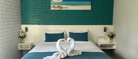 A hotel room with a bed, teal accent wall, white bedding, two nightstands with lamps, and a painting of a beach scene.