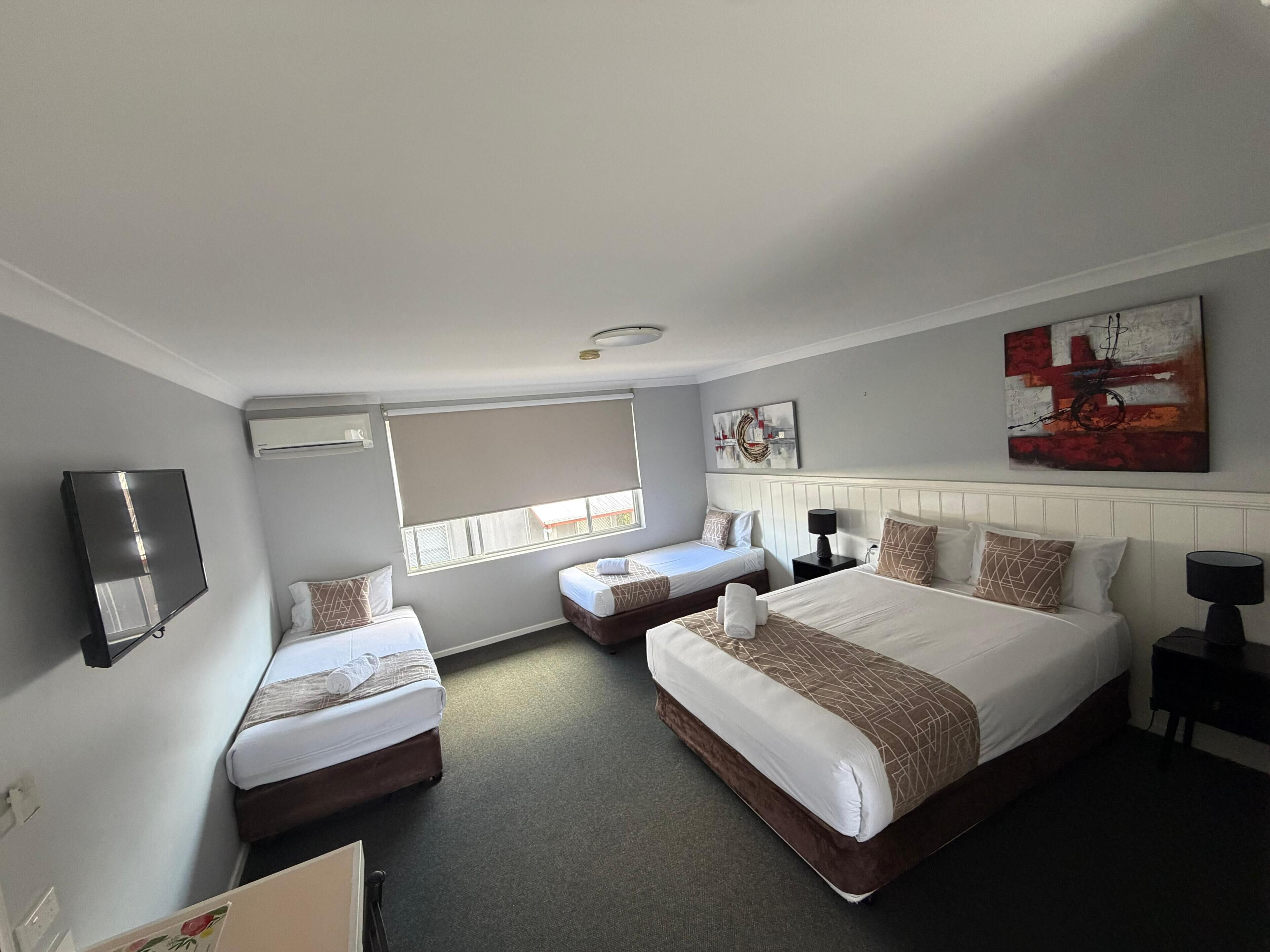 Ein Hotelzimmer mit zwei Einzelbetten, einem Kingsize-Bett, einem Flachbildfernseher, einem Fenster mit Jalousien und einem Bild an der Wand.