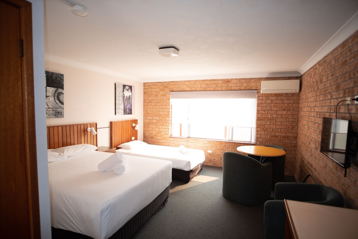 A hotel room with two beds, a small table, a chair, a window, and a brick wall.