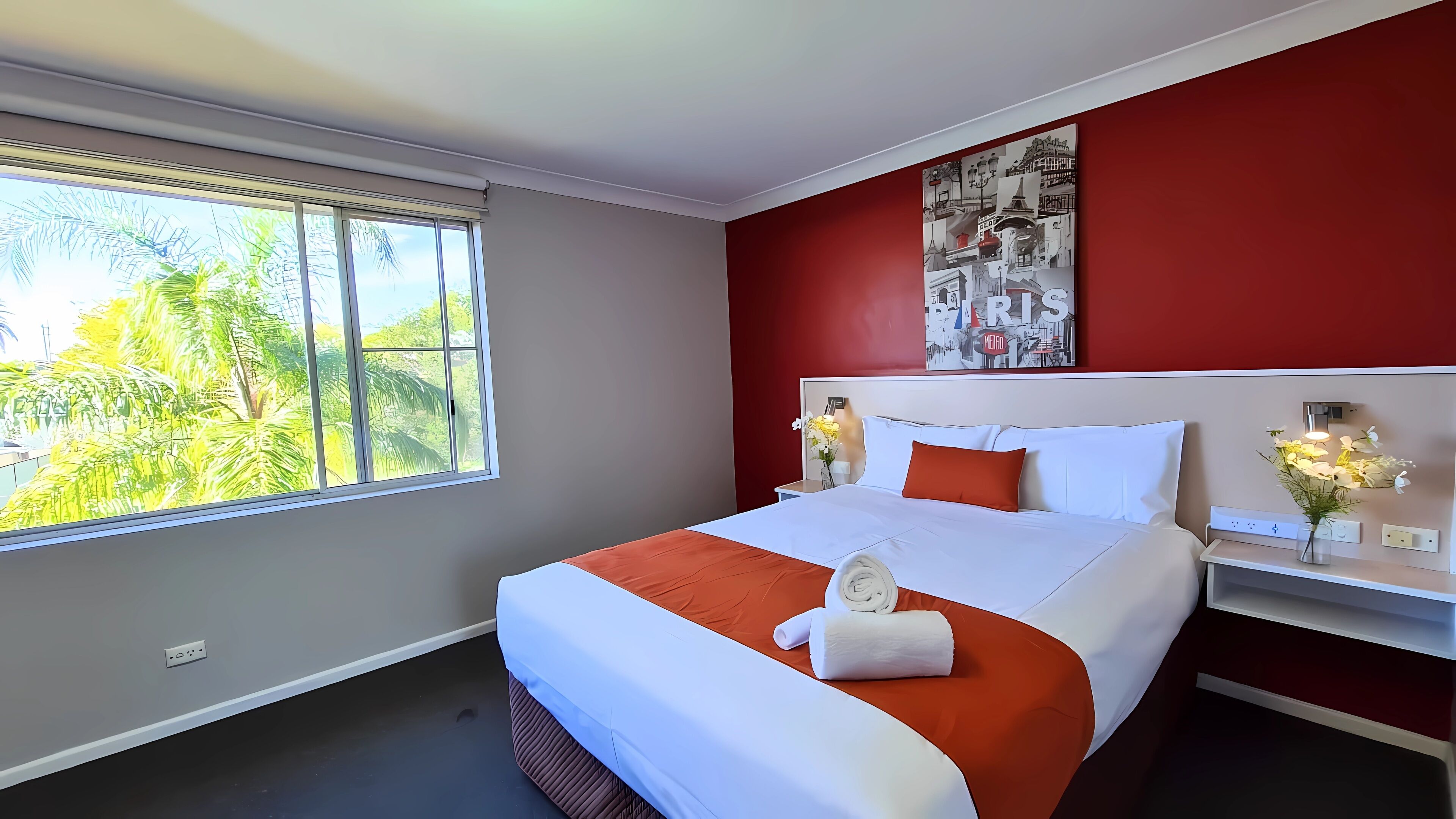 A hotel room with a large bed, a red accent wall, a window with a view of greenery, and a Paris-themed artwork.