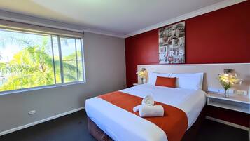A hotel room with a large bed, a red accent wall, a window with a view of greenery, and a Paris-themed artwork.