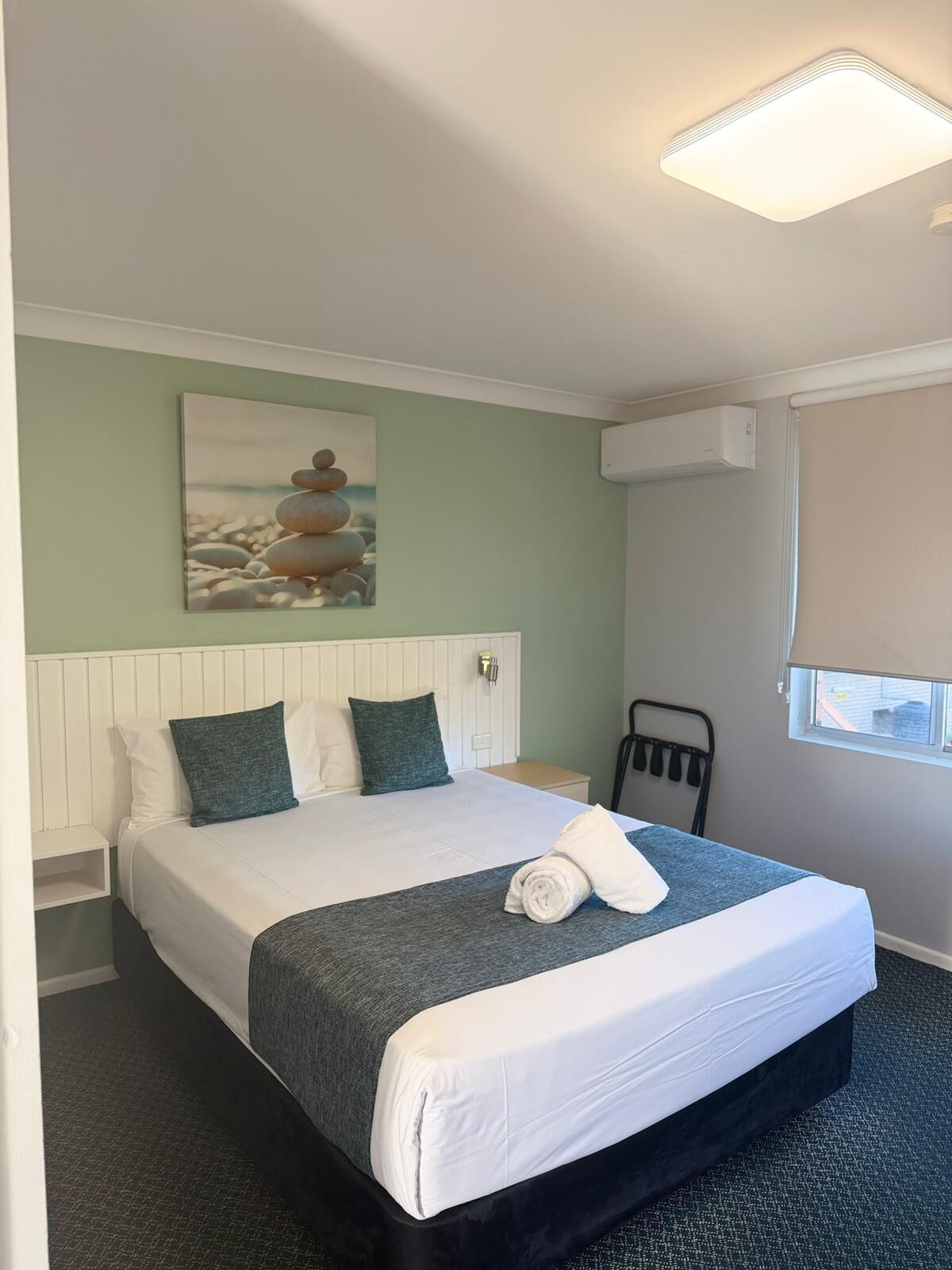 Una habitación de hotel con cama, un cuadro, equipo de aire acondicionado y una ventana con vistas.