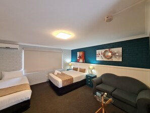 A hotel room with a bed, a sofa, a glass coffee table with a phone, and artwork on the wall.
