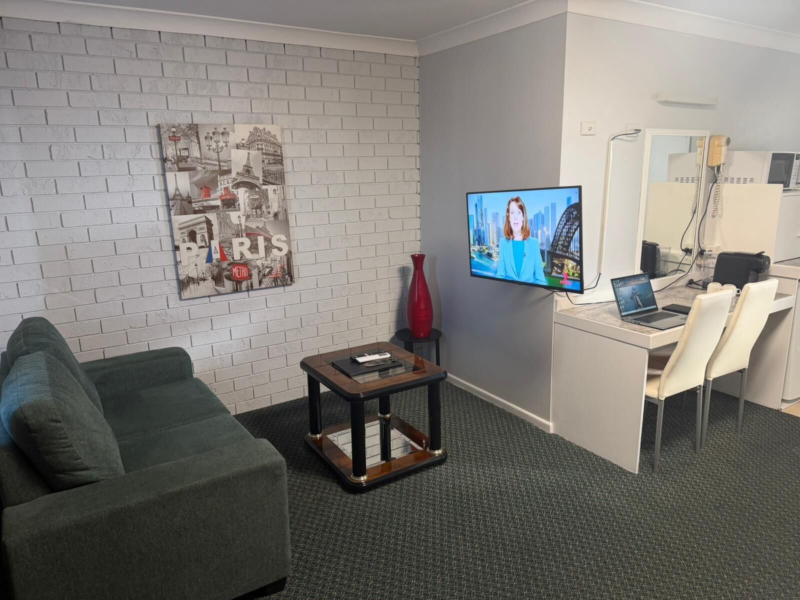 Quarto de hotel moderno com parede de tijolos cinza, TV de tela plana, escrivaninha com laptop, um vaso vermelho e um sofá verde.