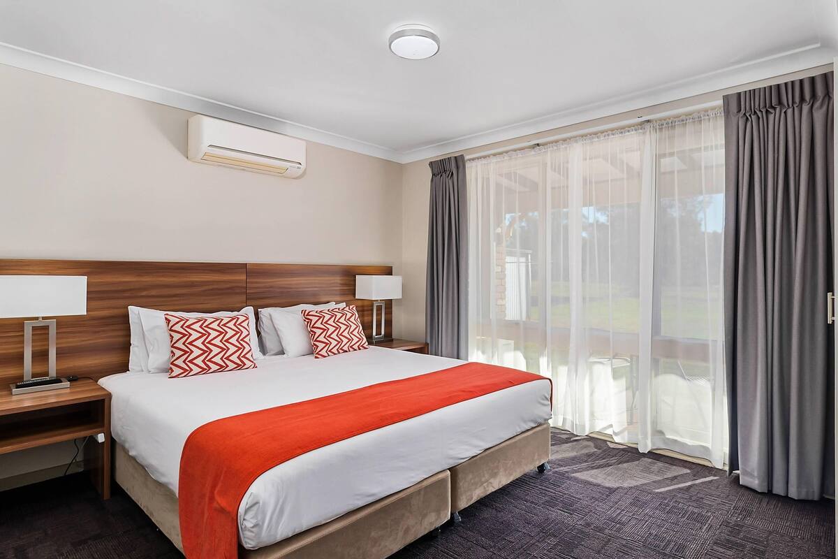 A hotel room with a large bed, two bedside tables with lamps, a ceiling air conditioner, and a view through a window with curtains.