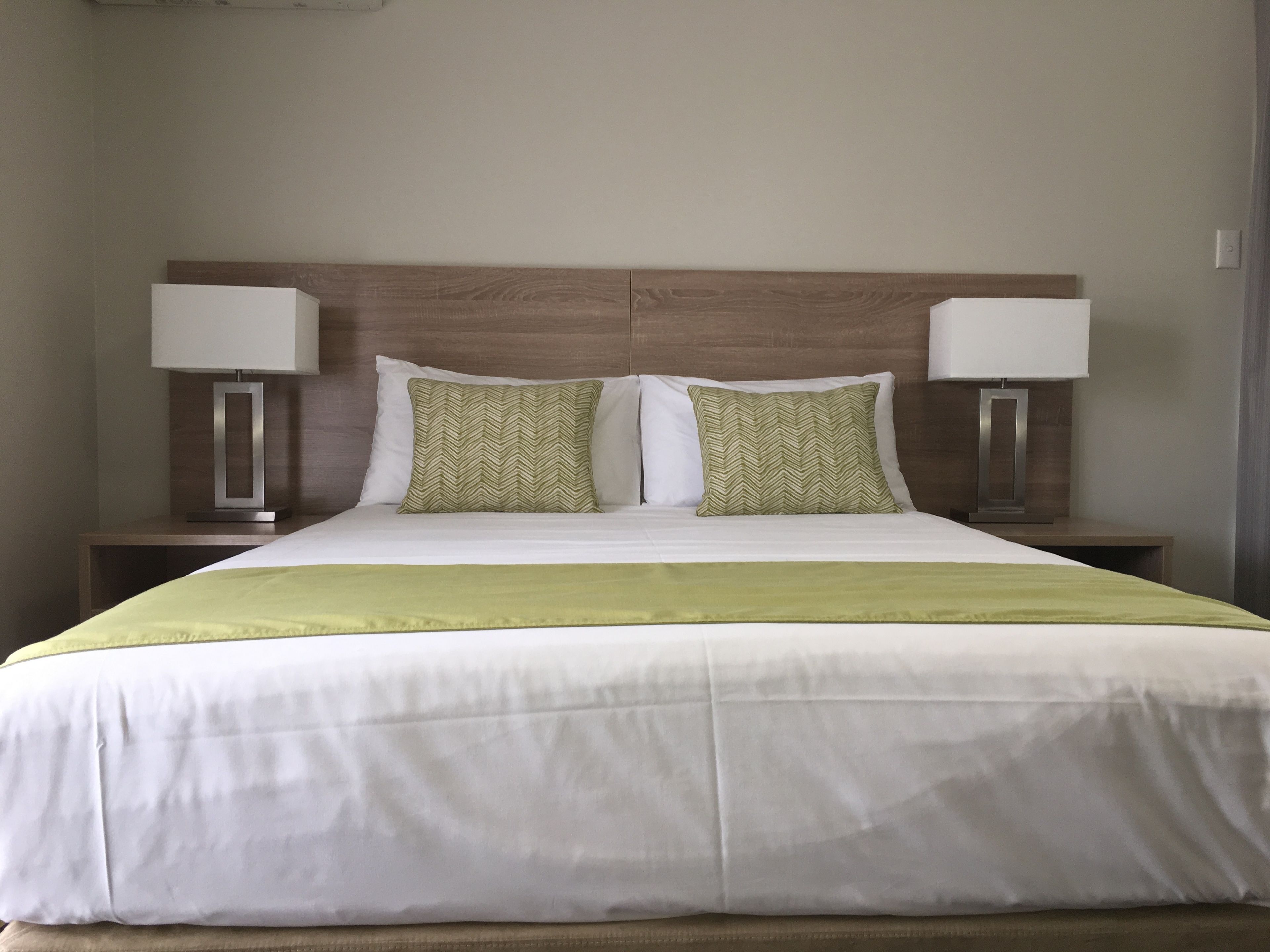 A neatly made bed with a green and white bedspread, two pillows with a green pattern, and two bedside lamps on wooden nightstands.