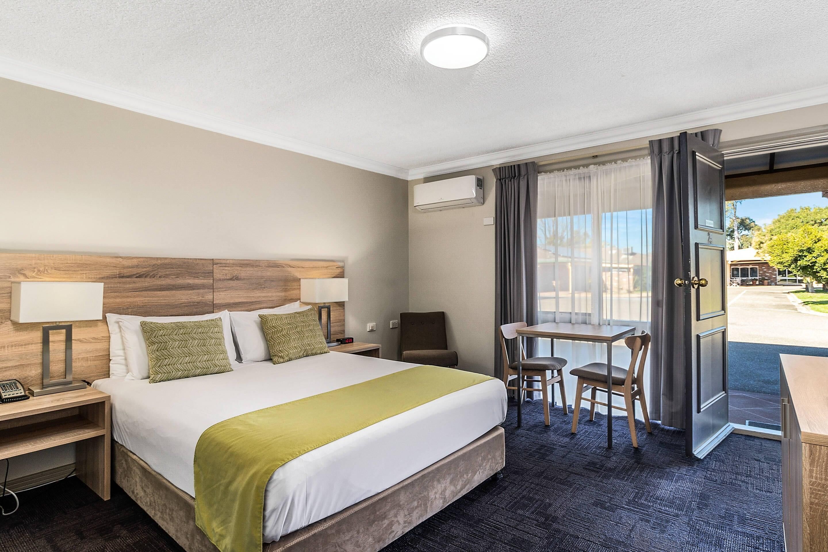 A hotel room with a bed, bedside lamps, a desk with chairs, and a view through an open door.