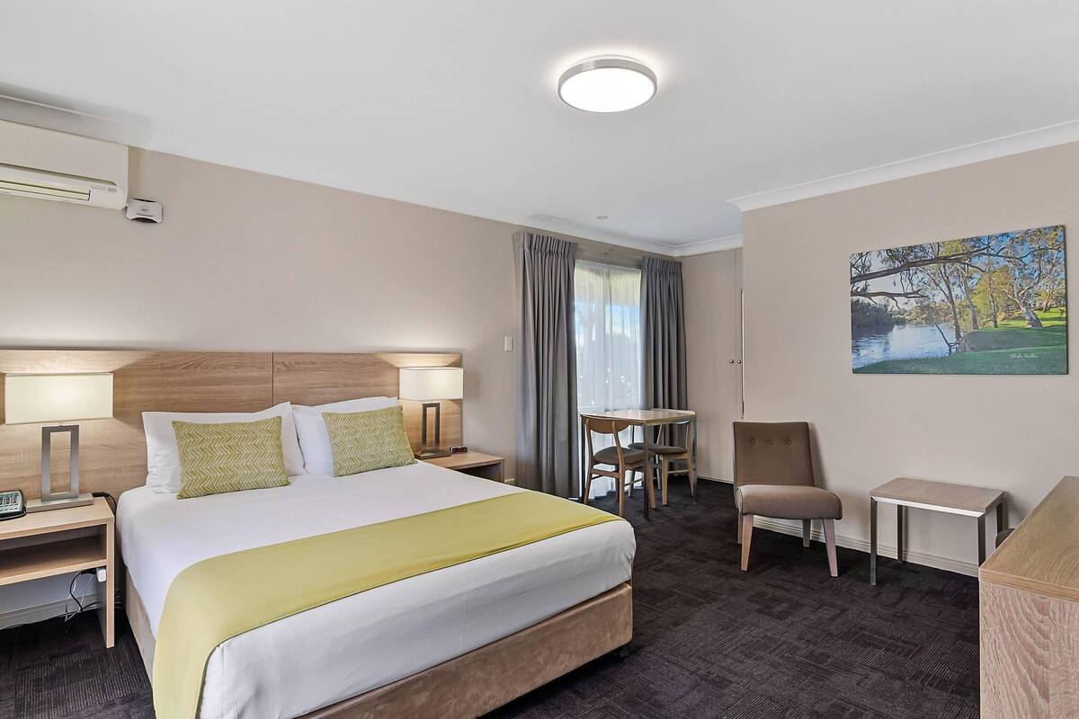 A hotel room with a large bed, bedside lamps, a desk, a chair, and a painting on the wall.