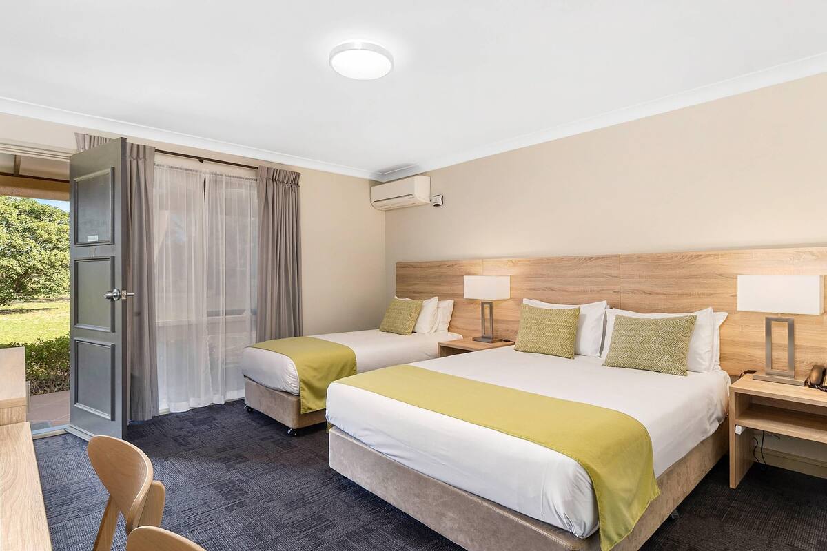A hotel room with two beds, a desk, a chair, an air conditioning unit, and a view of the outdoors.