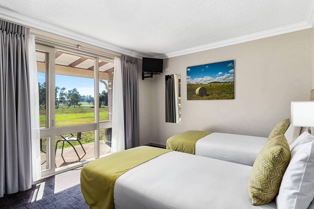 A hotel room with two beds, a large window with curtains, a painting on the wall, and a view of the outdoors.