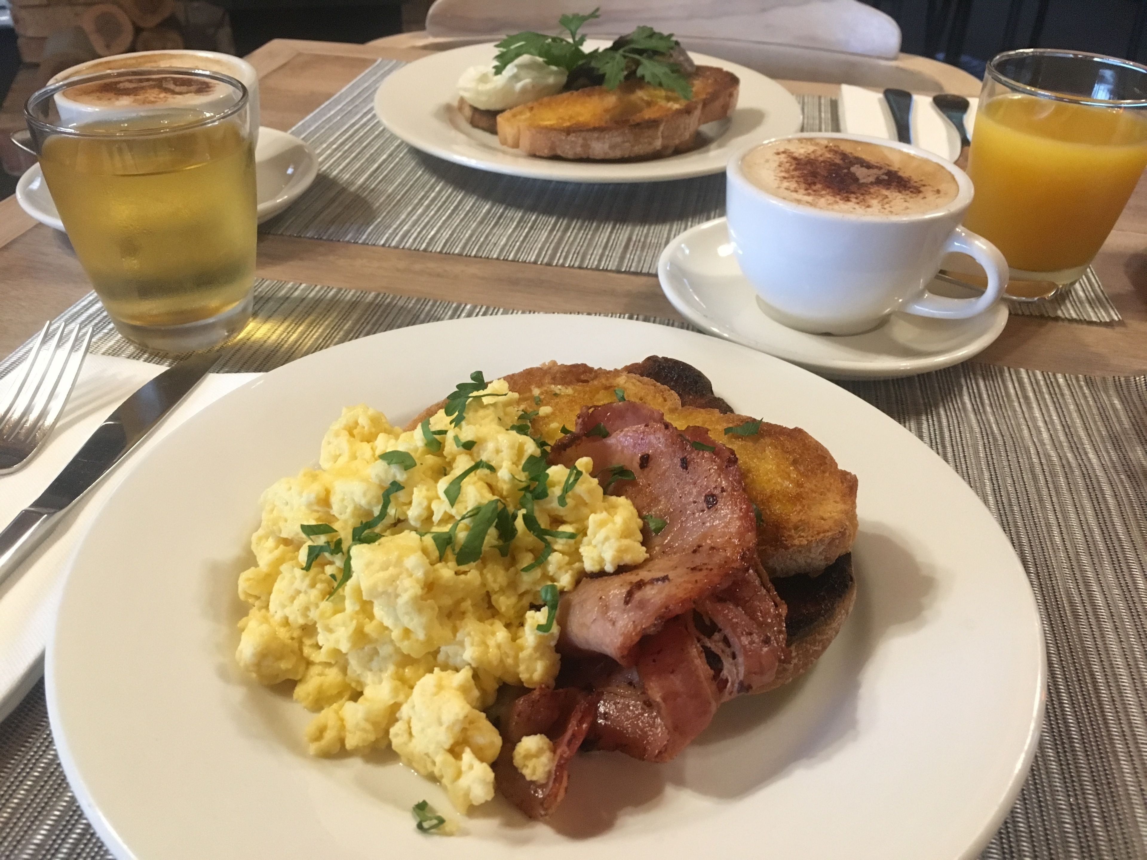 Desayuno con huevos revueltos, tocino, tostadas y bebidas sobre una mesa.