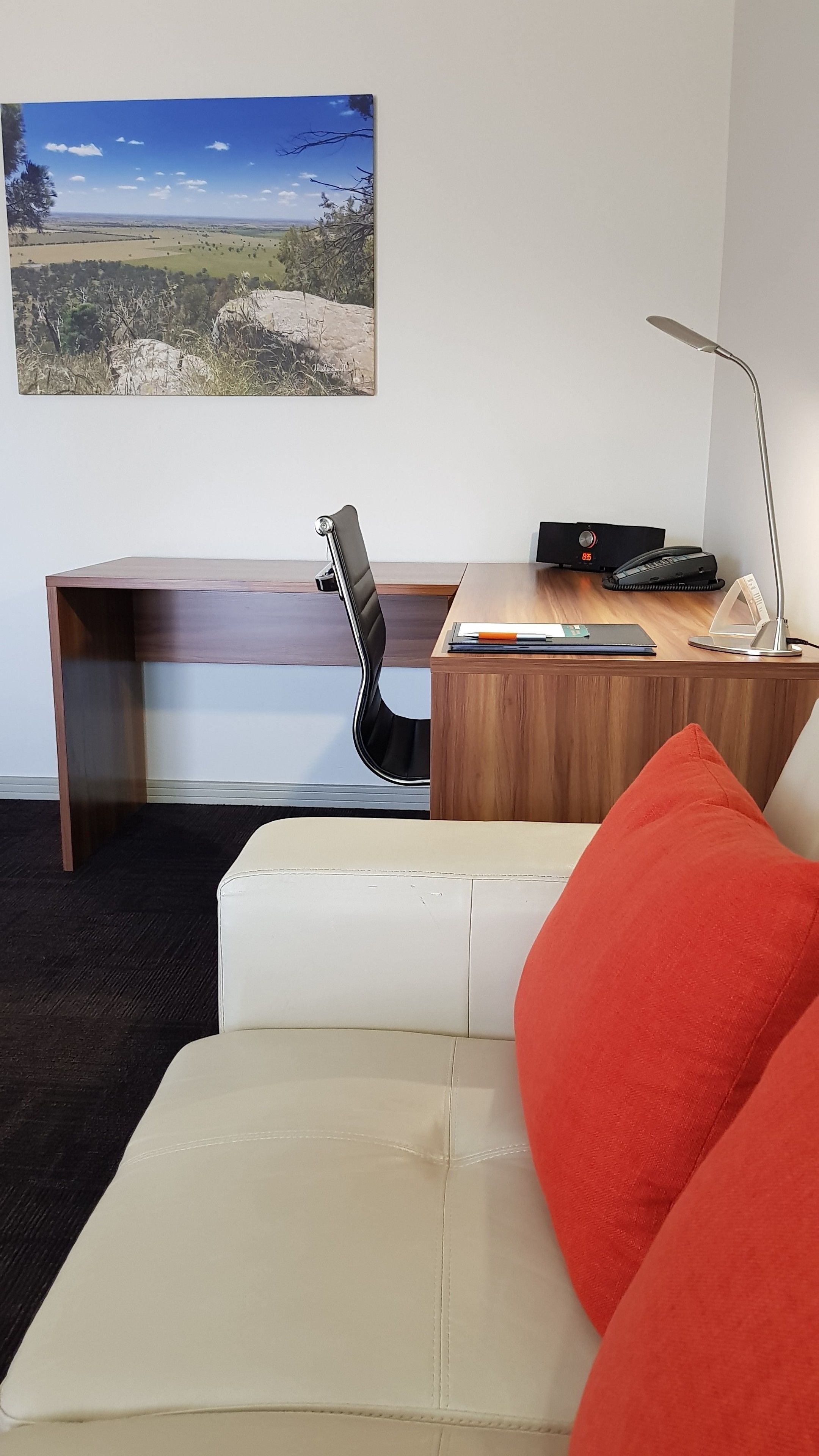 Una habitación moderna con un escritorio de madera, una silla de oficina negra, un sofá blanco con un cojín rojo y un cuadro de paisaje en la pared.