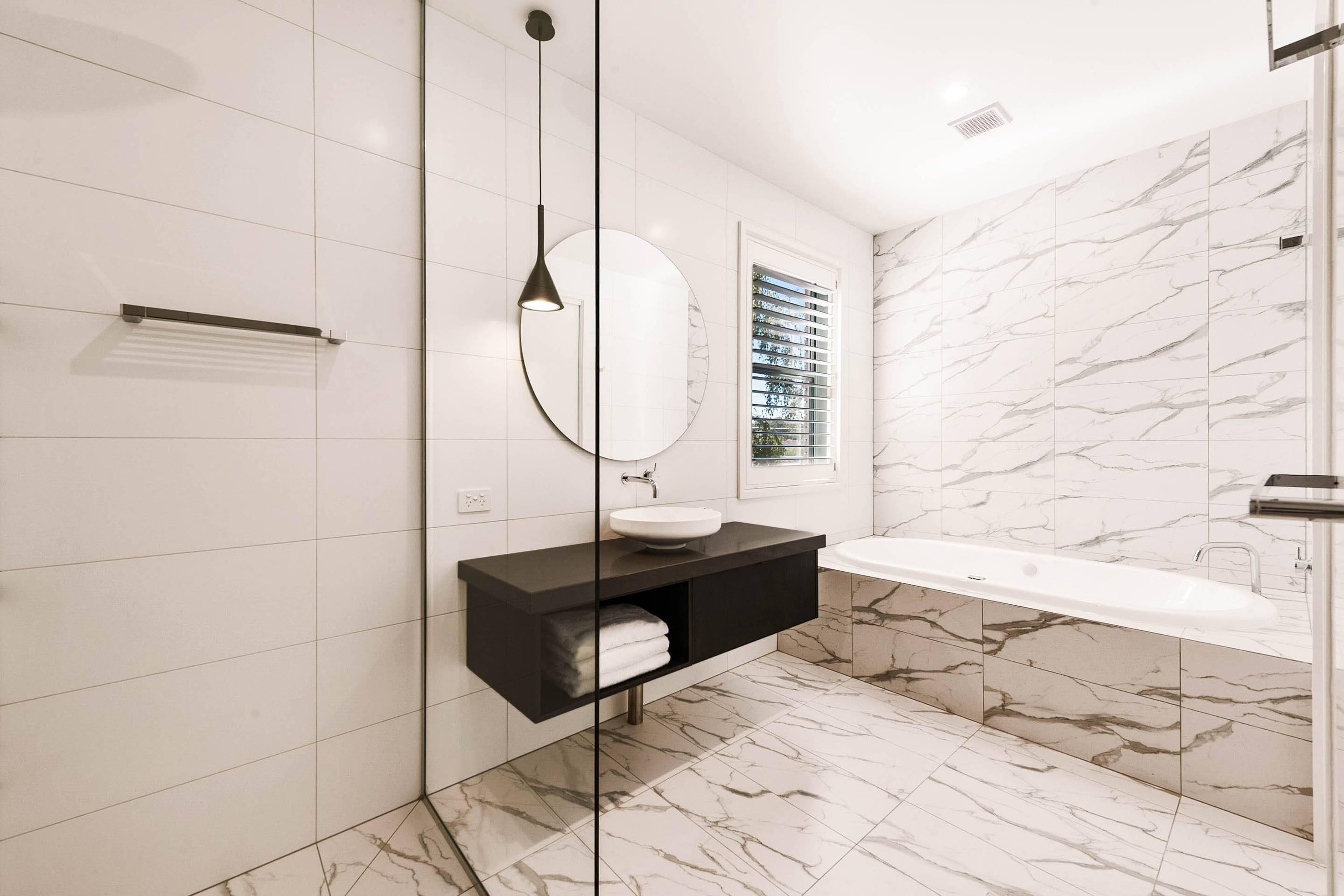 Un baño moderno con piso de mármol, un mueble de tocador negro con espejo redondo, un lavamanos blanco y una bañera independiente.
