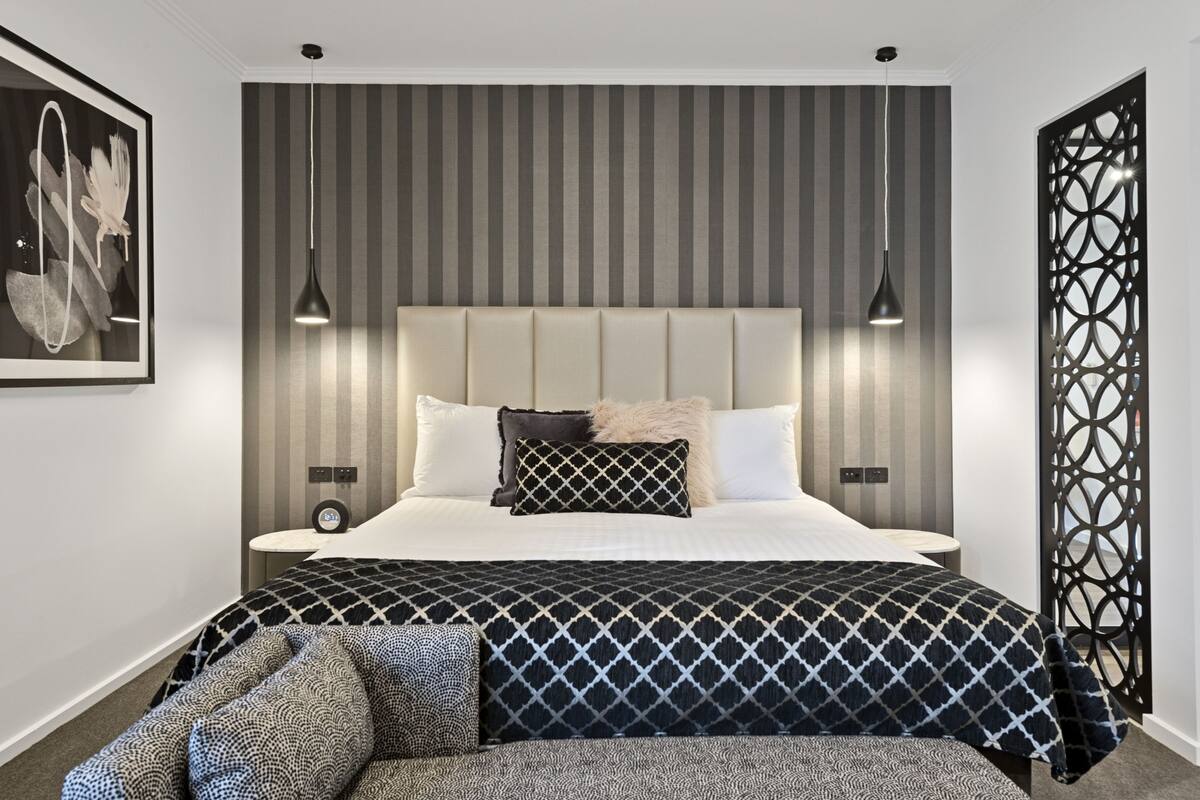A modern bedroom with a large bed, striped wallpaper, two pendant lights, and a decorative wall panel.