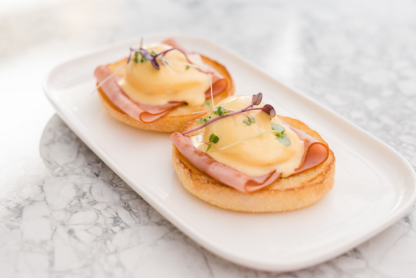 Twee Engelse muffins belegd met ham en Hollandaisesaus, gegarneerd met microgroenten, op een wit rechthoekig bord.
