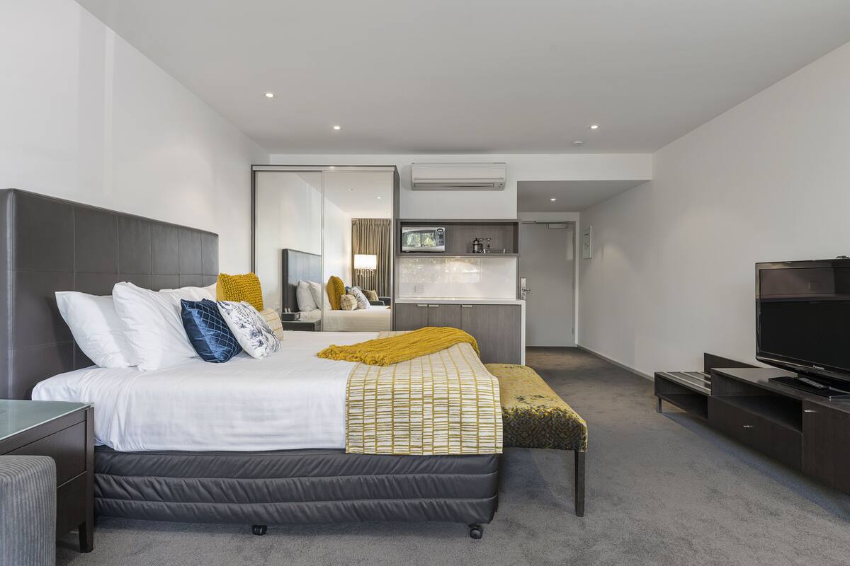 A modern hotel room with a large bed, a TV, a desk, and a bathroom visible through an open door.