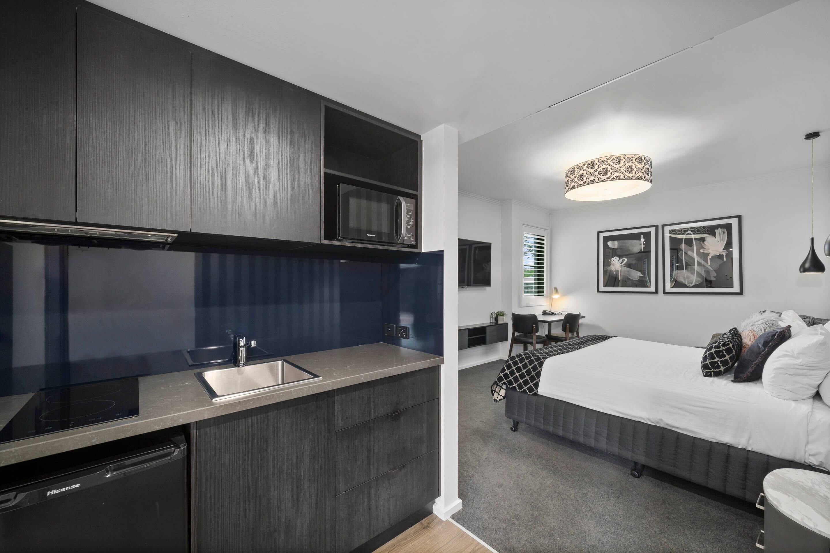 A compact hotel room with a kitchenette, a bed, a bedside table, and a wall-mounted lamp.