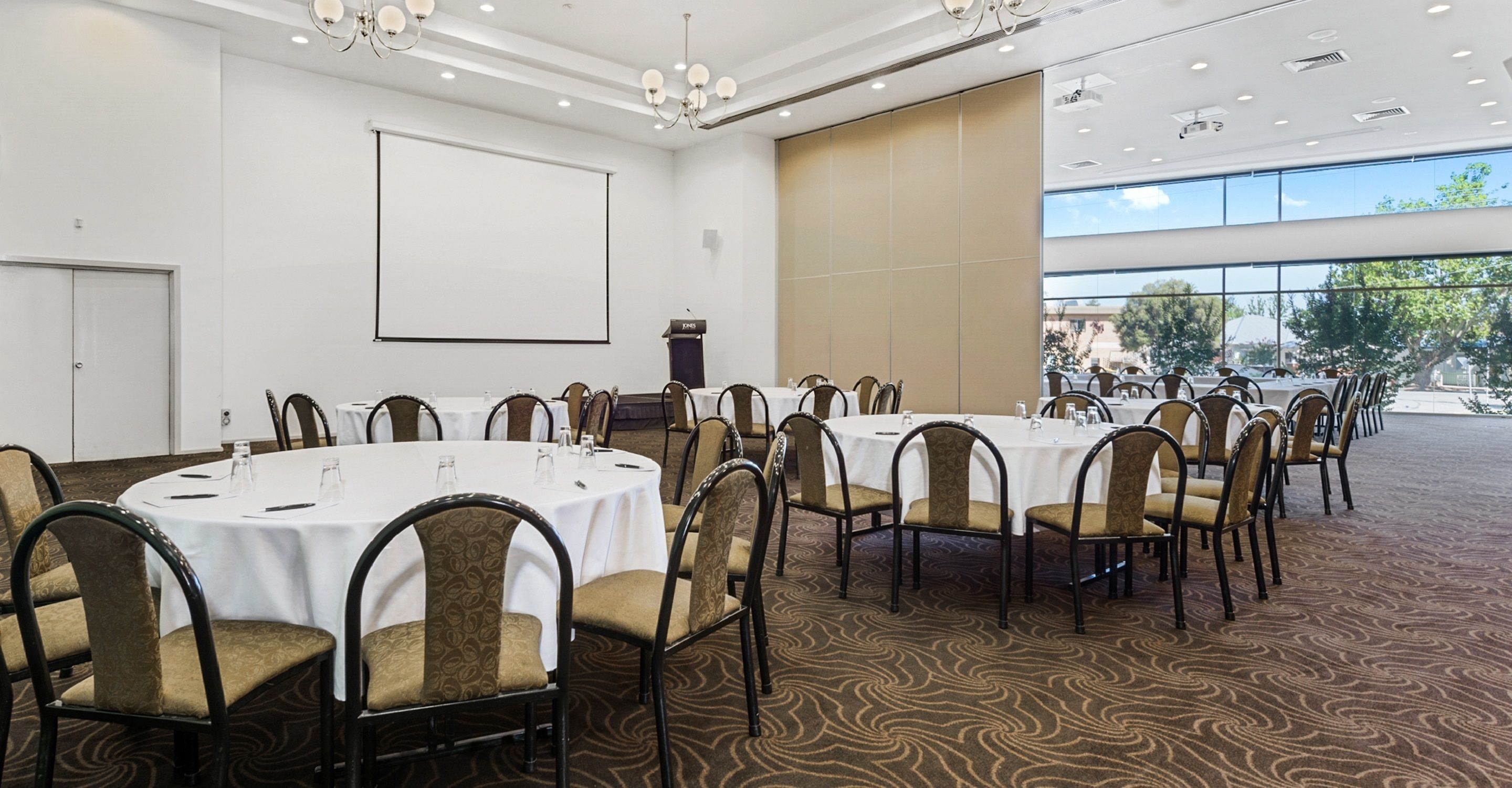 Un salón de conferencias con mesas redondas, sillas, una pantalla de proyector y un atril.