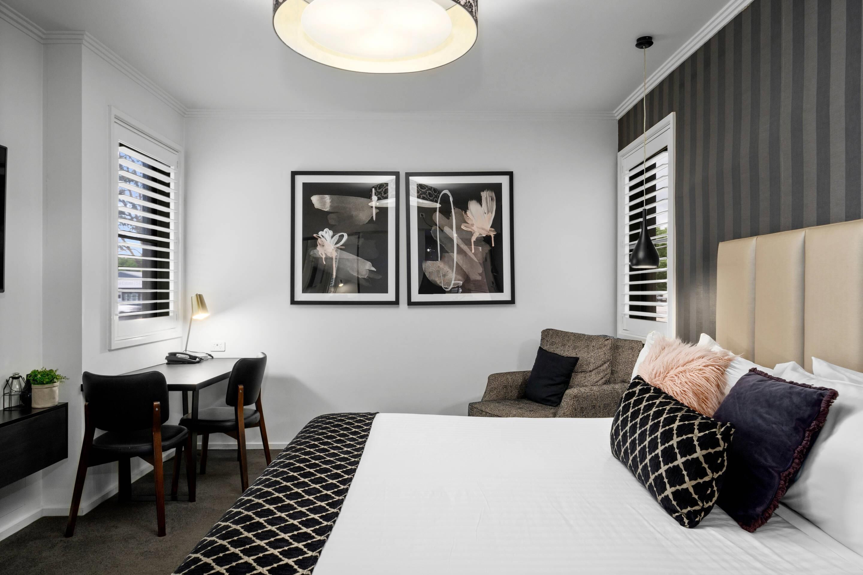 A modern hotel room with a large bed, a desk, two framed pictures on the wall, and a window with blinds.