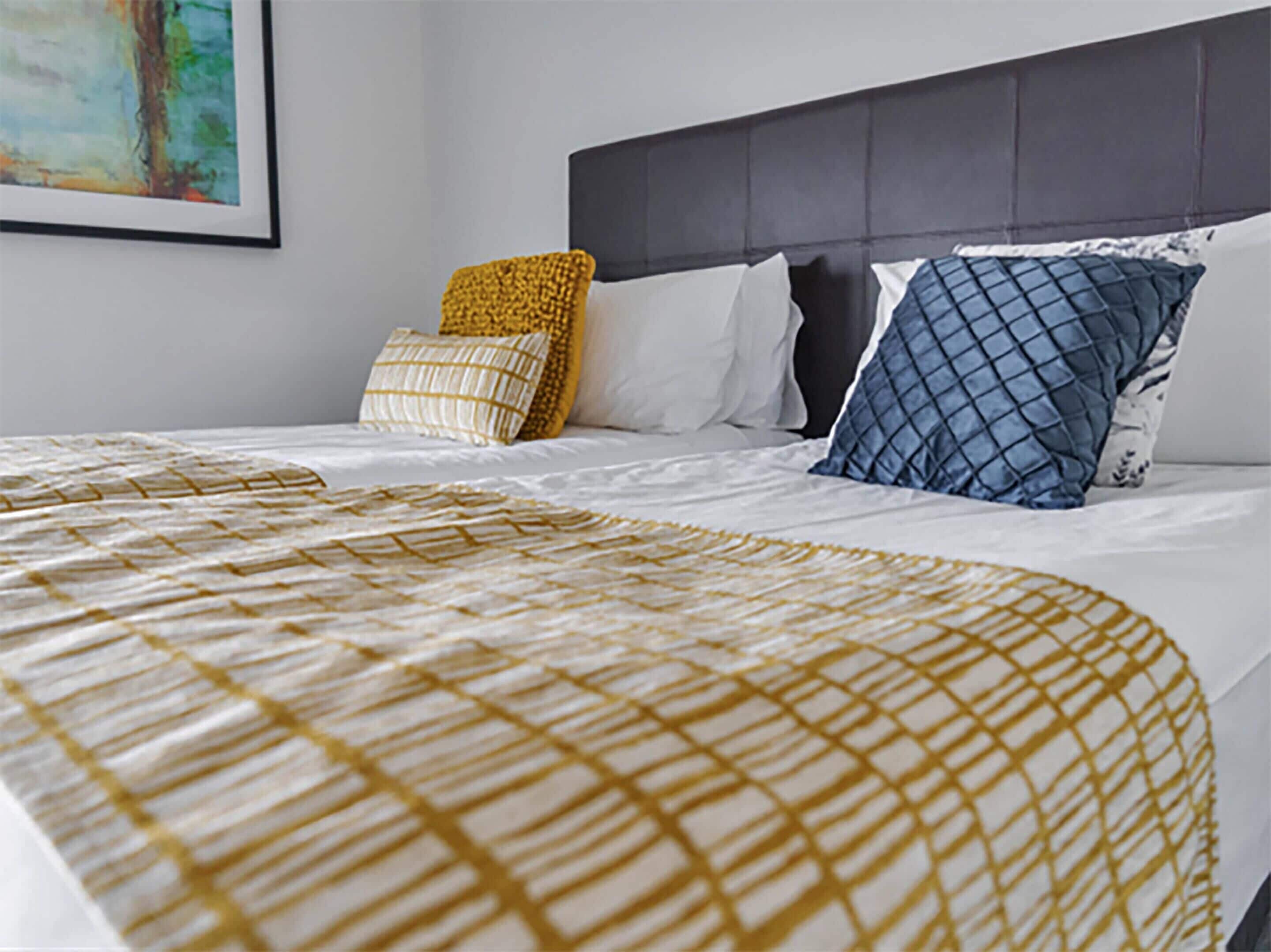 Una cama bien hecha con una colcha estampada, un cabecero oscuro y varias almohadas con diferentes diseños y colores.