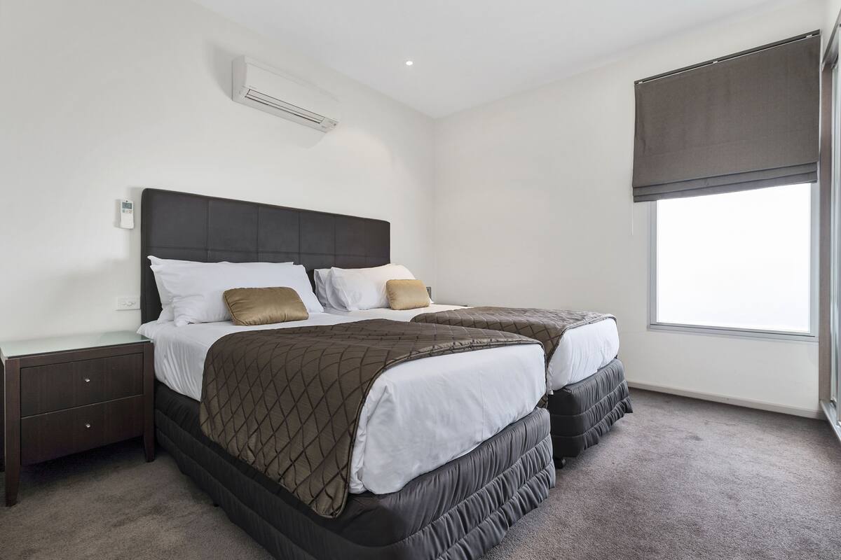 A bedroom with two beds, a nightstand, a wall-mounted air conditioner, and a window with a blind.
