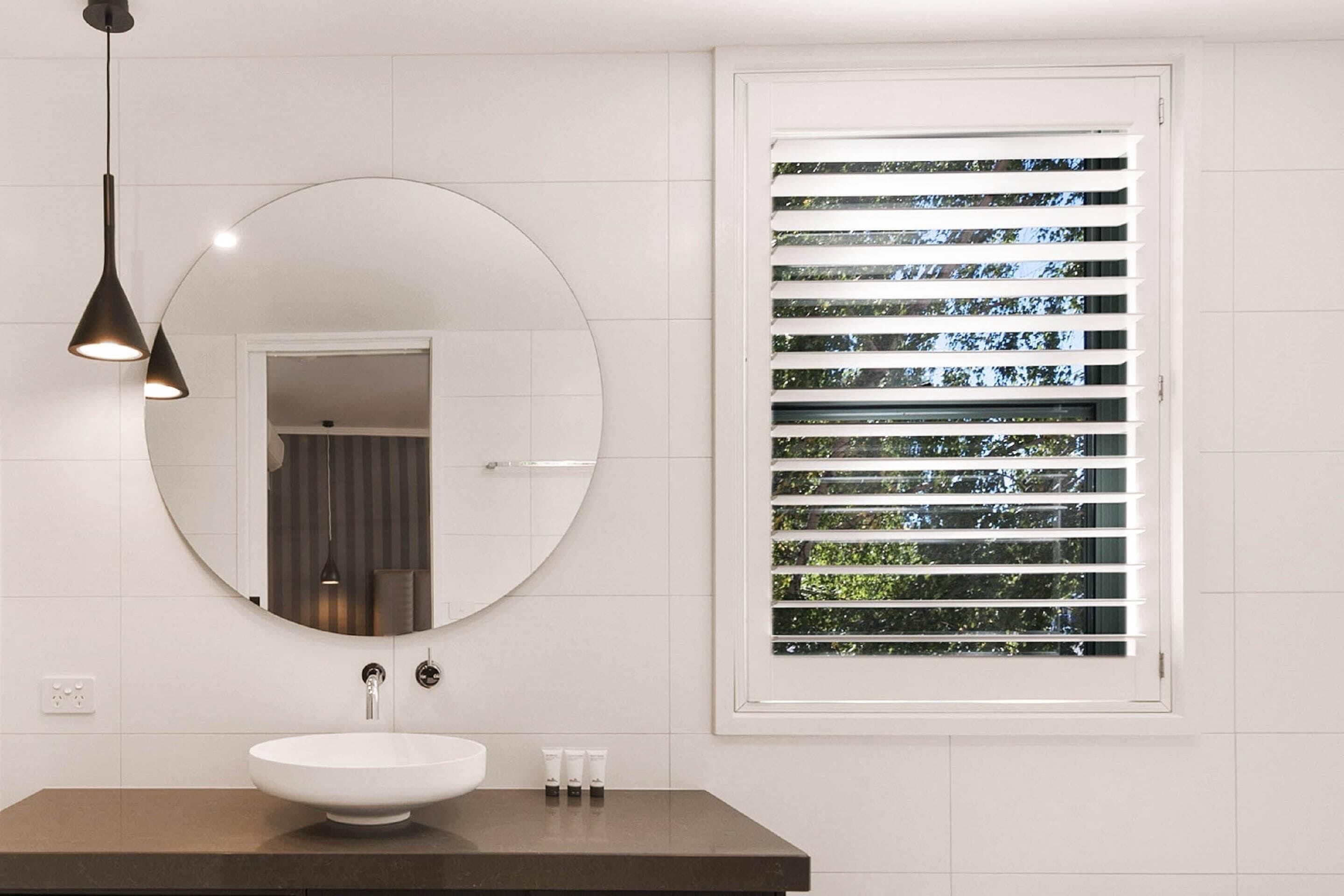 A bathroom with a round mirror, a white sink, and a window with blinds.