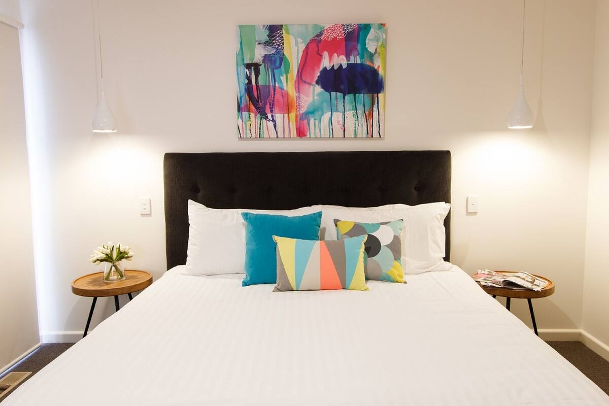 A hotel room with a large bed, two bedside tables, a wall-mounted abstract painting, and pendant lighting.