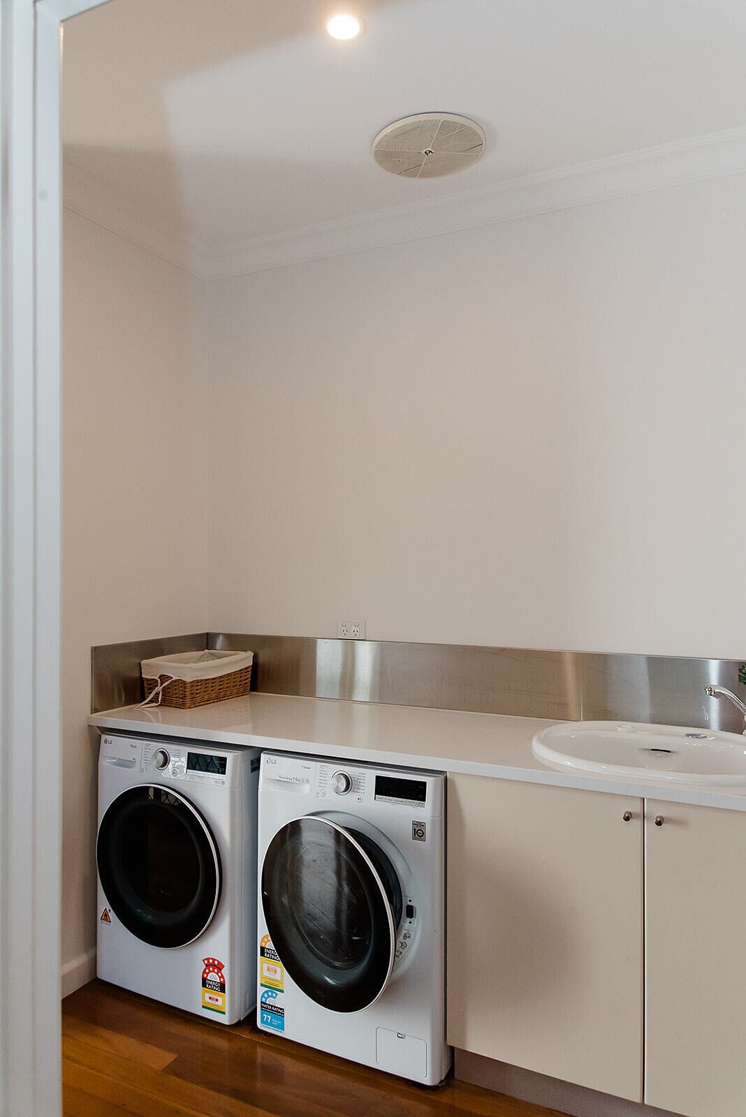 A laundry room with two front-loading washing machines, a stainless steel sink, and a basket on the countertop.