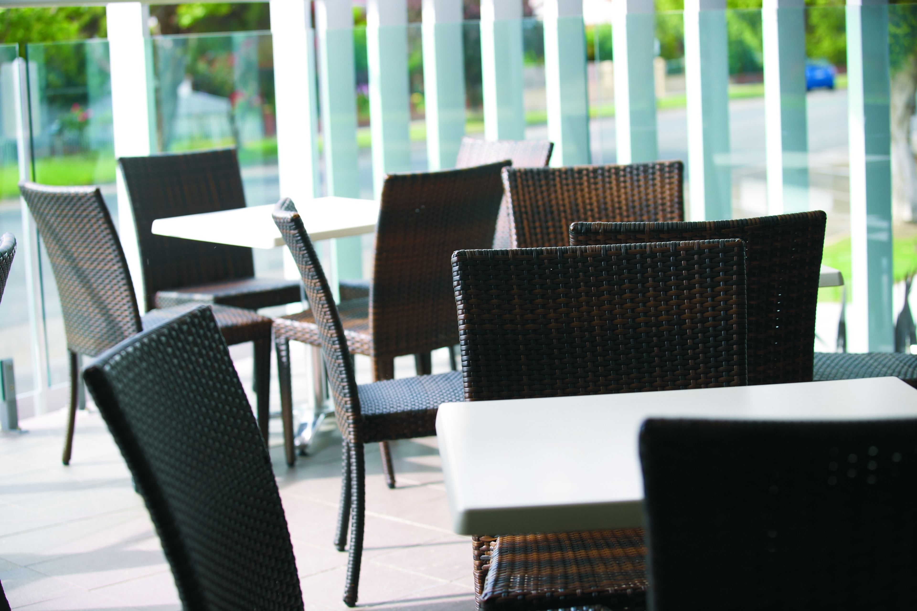 Des chaises et des tables en osier sur une terrasse extérieure, avec une rambarde en verre et vue sur la rue.