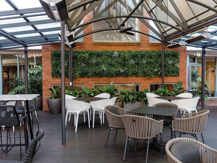 An outdoor seating area with tables and chairs, a brick wall with greenery, and a glass ceiling structure.