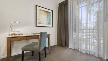 A room with a desk, chair, lamp, framed artwork, and large windows with curtains.