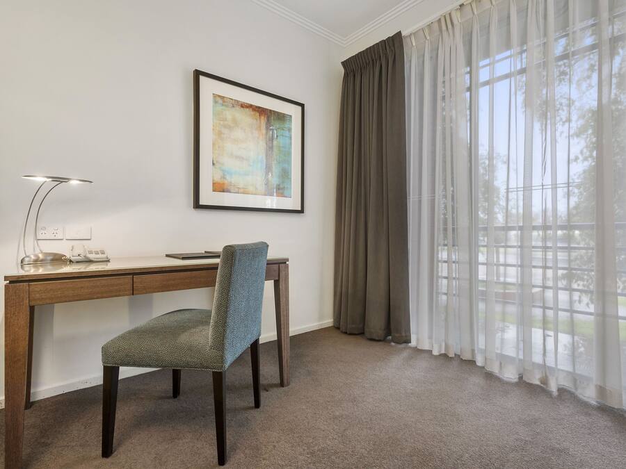 A room with a desk, chair, lamp, framed artwork, and large windows with curtains.
