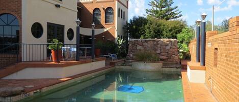 A swimming pool area with a stone wall, a lounge chair, and a building with arched windows.
