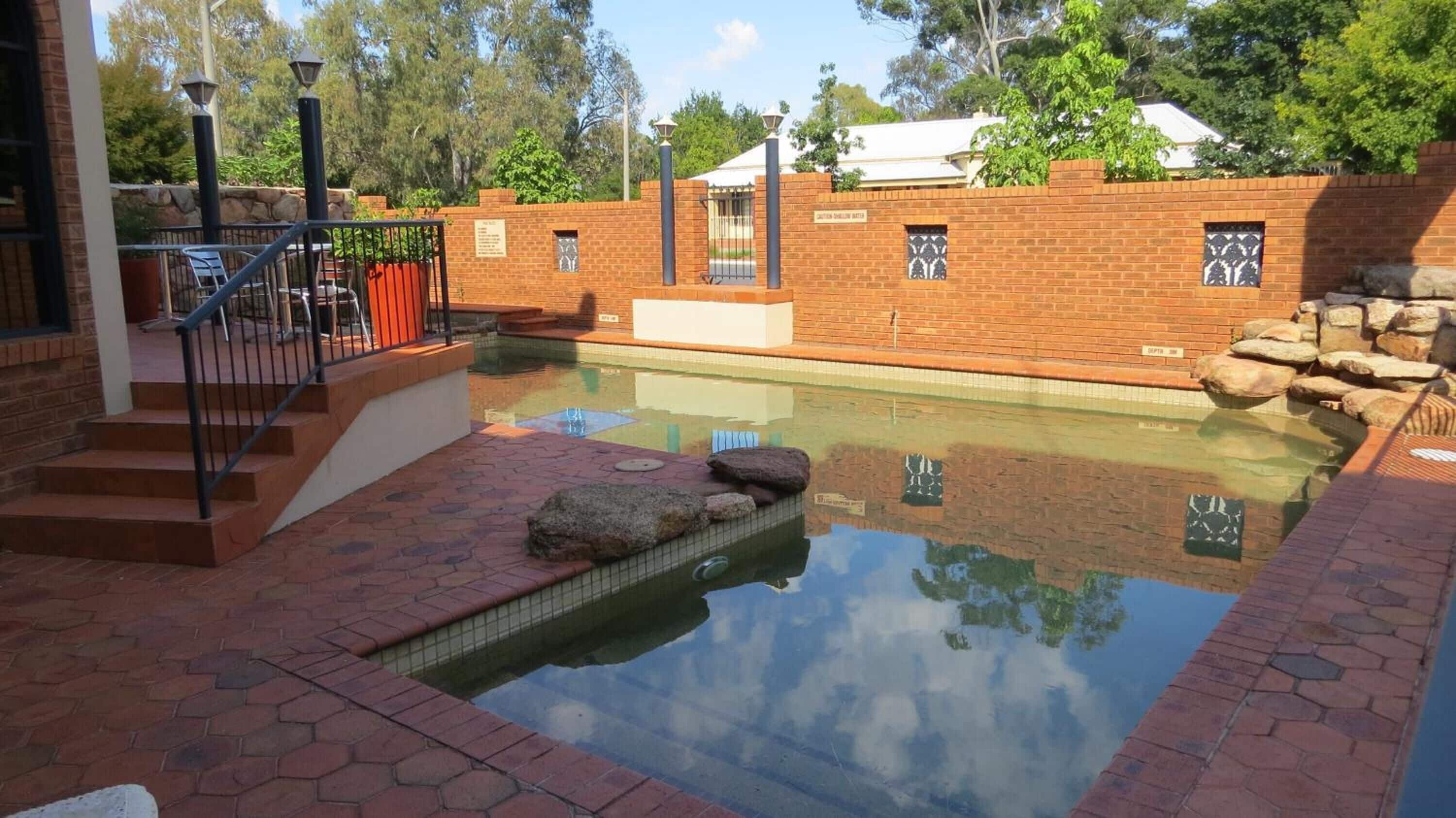 A swimming pool with a brick wall and a small bridge leading to a patio area.