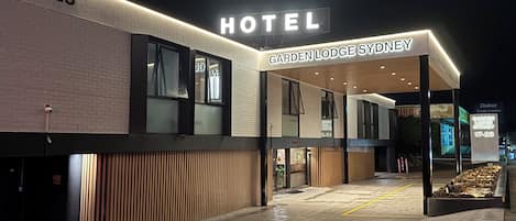 En modern hotellentréd med en skylt som visar texten ”HOTEL GARDEN LODGE SYDNEY” och en tydlig adress: ”17–23”.